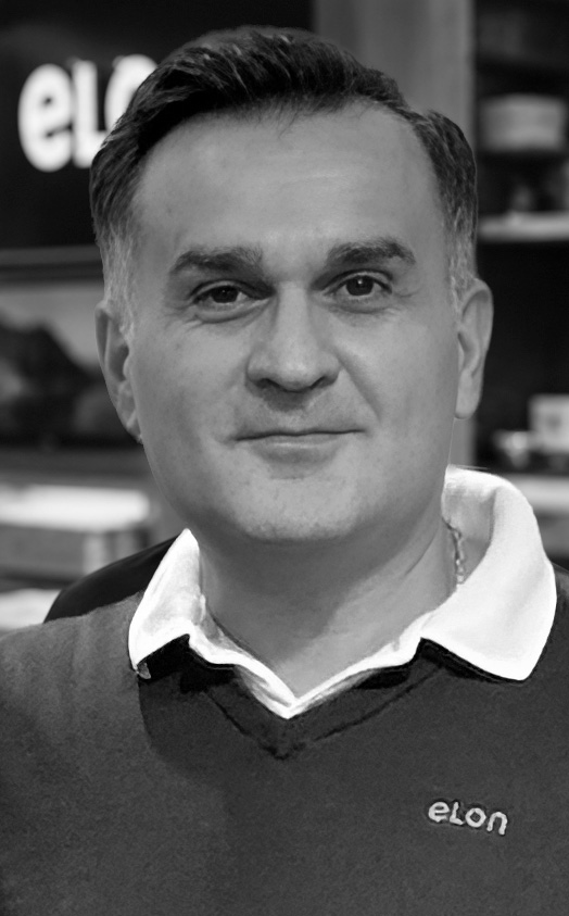 Portrait of a man with short dark hair, wearing a dark sweater with a white collared shirt, smiling at the camera indoors. Background is blurred with shelves visible.