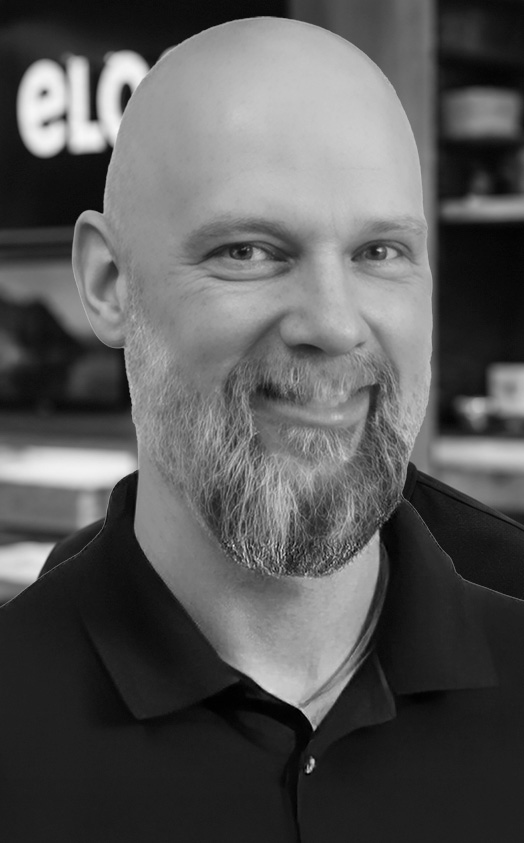 Portrait of a smiling bald man with a gray beard wearing a black polo shirt, in a professional headshot.